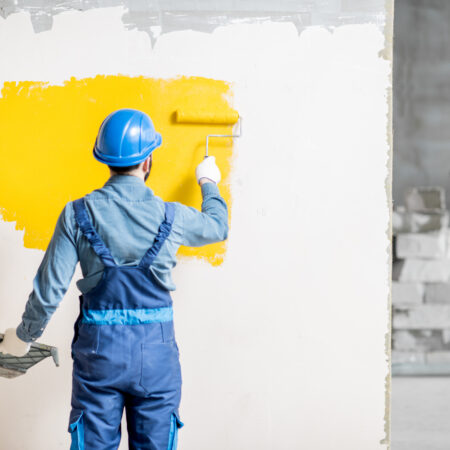 Workman in uniform painting wall with yellow paint at the construction site indoors